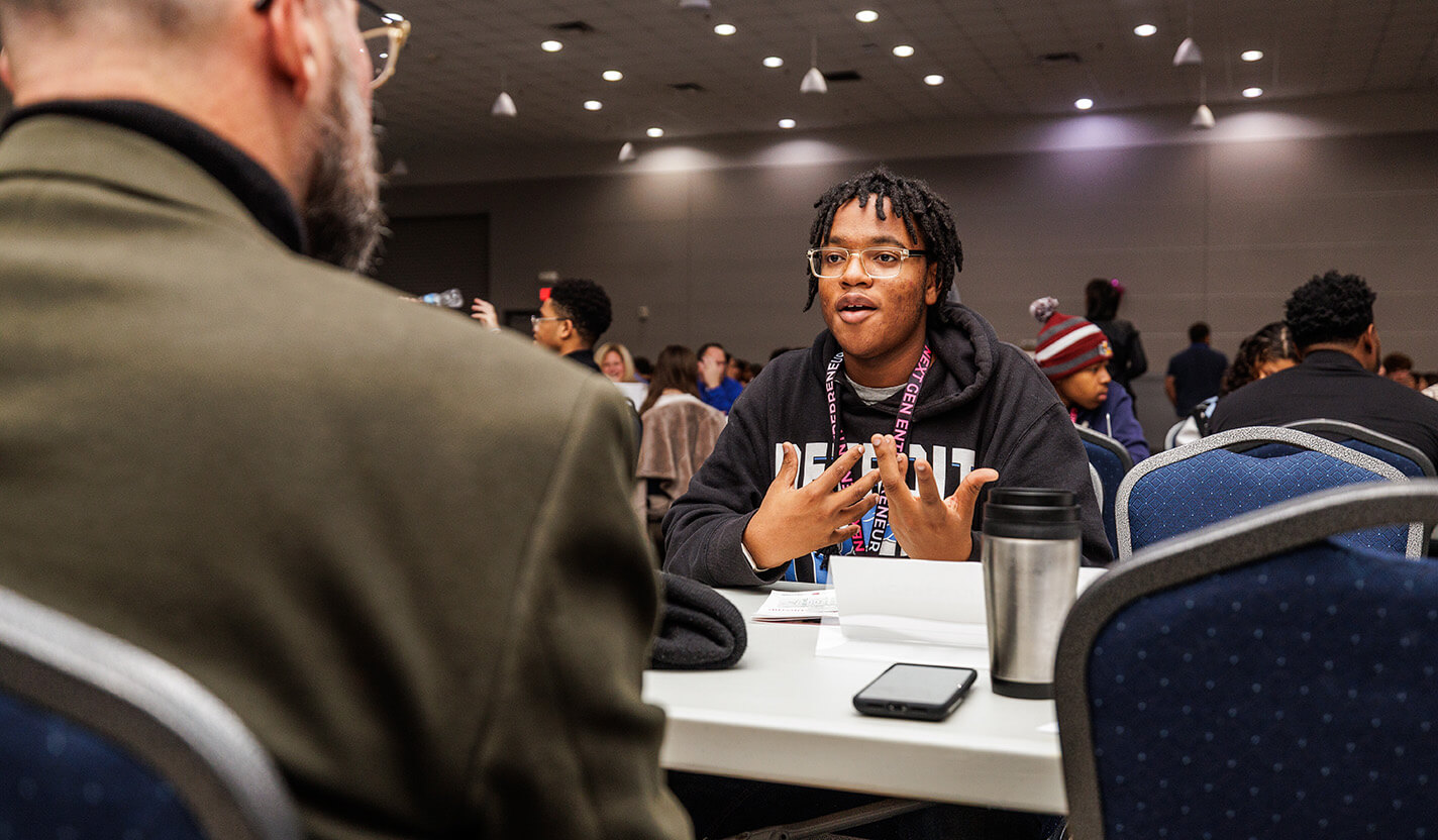 Teen in a black sweatshirt speaks to an adult at a table.