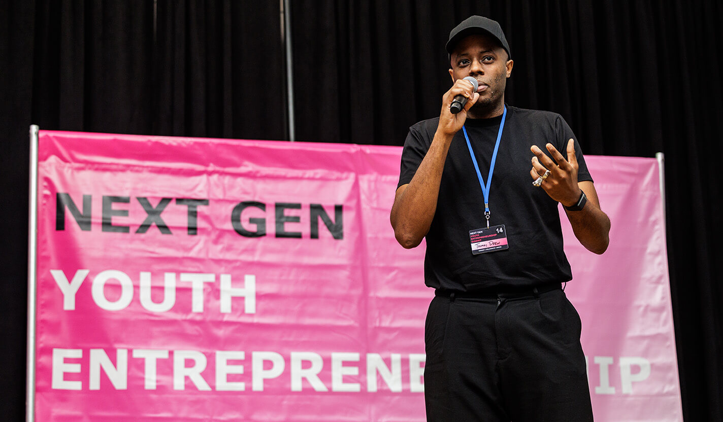 Man in a black T-shirt and cap speaks into a microphone in front of a pink banner that says, “NEXT GEN YOUTH ENTREPRENEURSHIP SUMMIT”