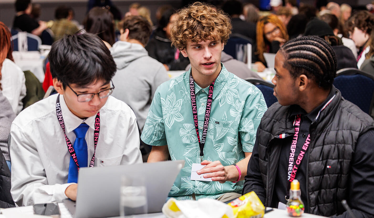 Three teen students in collared shirts sit talking to each other at a table.