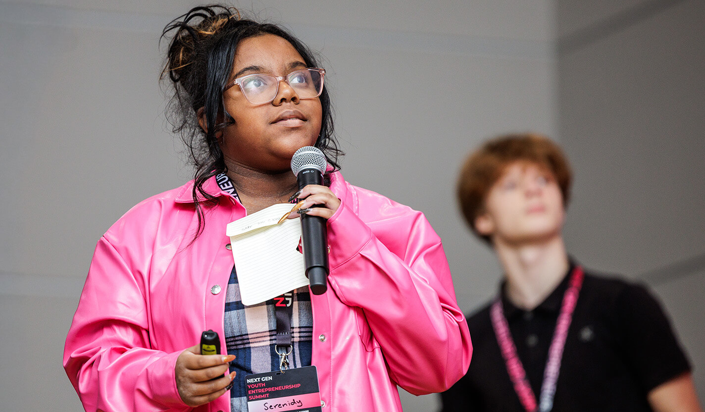Young woman in a pink jacket speaks into a microphone.