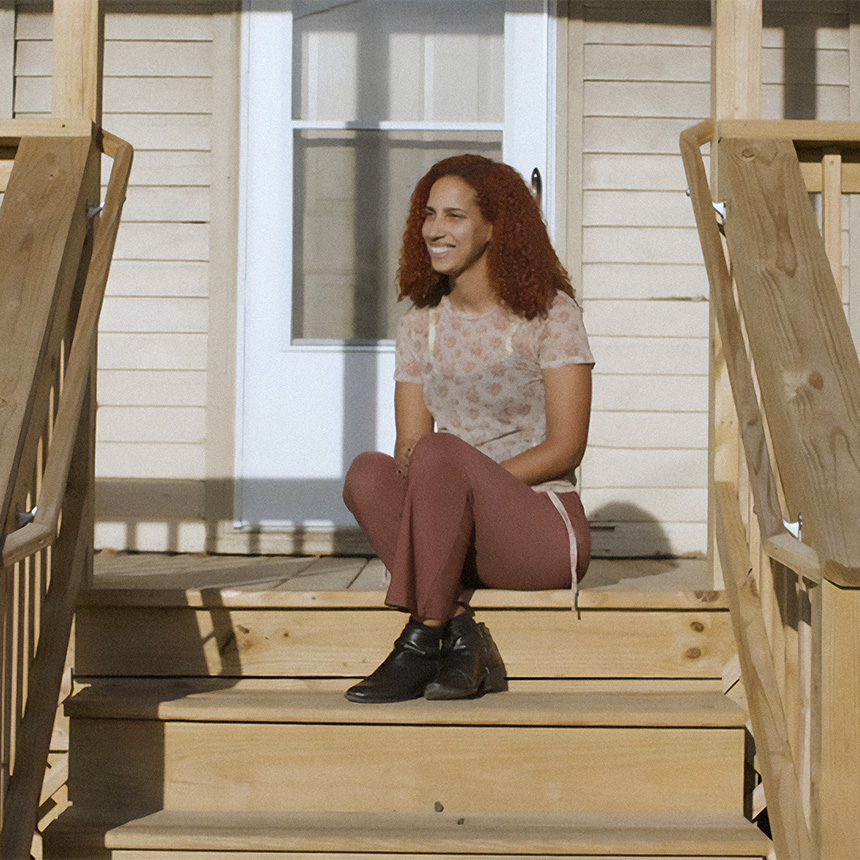 A young woman sits on. the steps of her newly build front porch as she smiles happily.