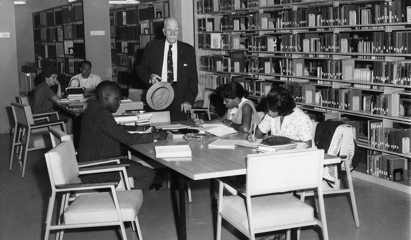 A man stands in the middle of the photo with young men and women sitting at two tables around him.