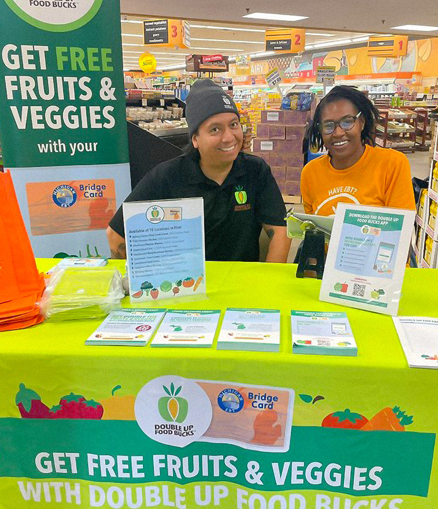 Two people sity at a table with signs around the table that advertise the Double Up Food Bucks benefits of getting free and reduced cost fresh fruits and vegetables.
