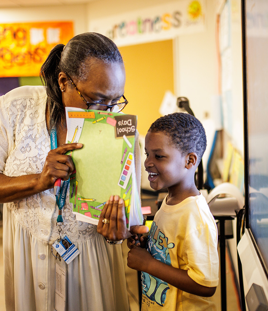 A teacher talks with a young boy in a classroom while holing up a stack of papers in front of them.
