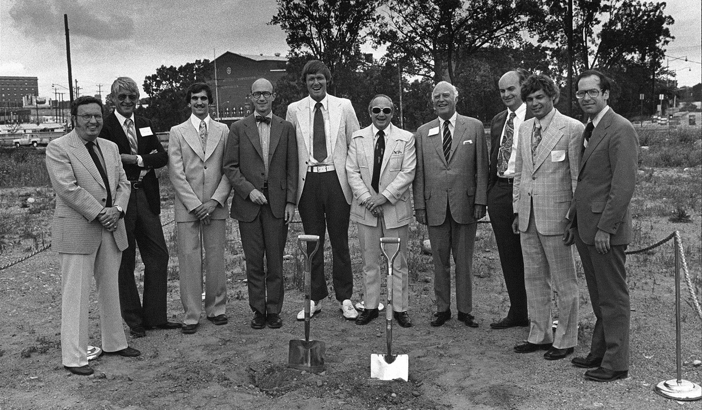 Ten men stand in a line with shovel in the dirt in front of them.