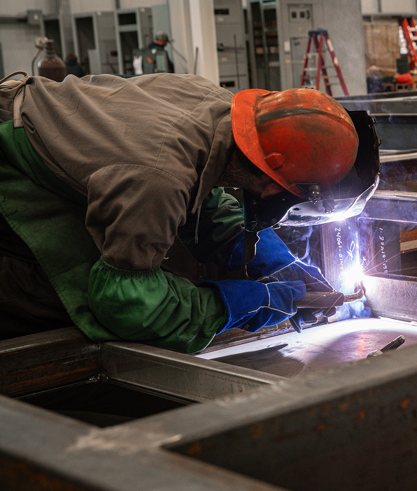 A man wearing an orange hard hat and a welding helmet bends over to weld pieces of metal.