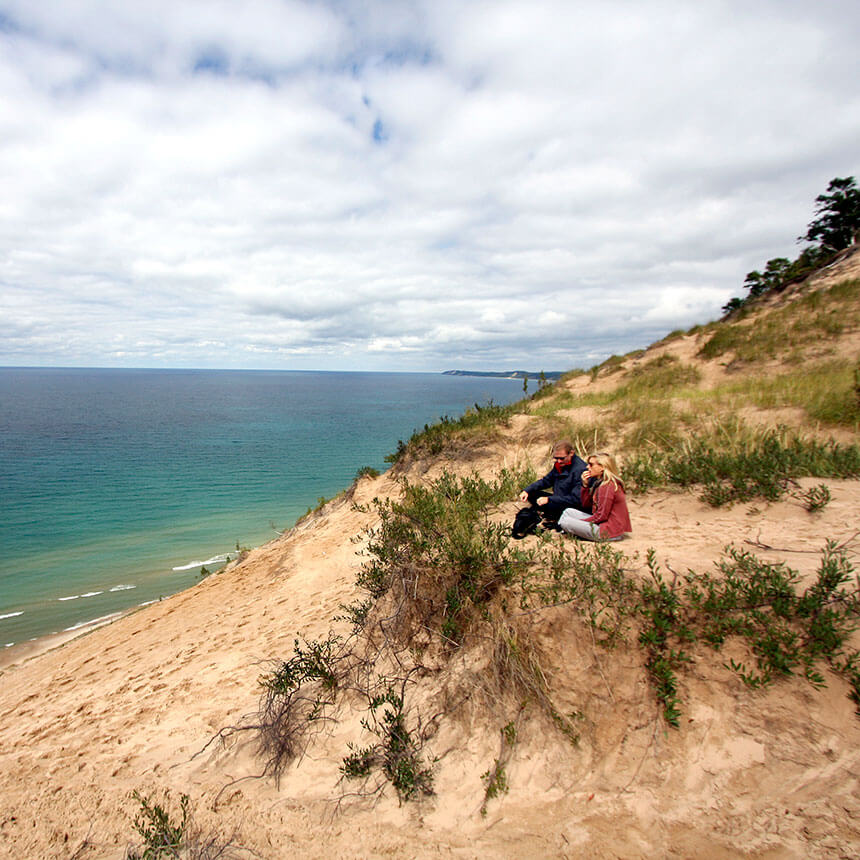 A couple sit atop a sandy dune and look out over a lake.