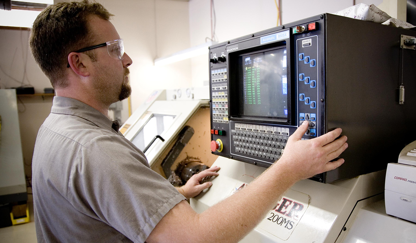 A man wearing safety glasses looks at a screen while pushing buttons on a machine.