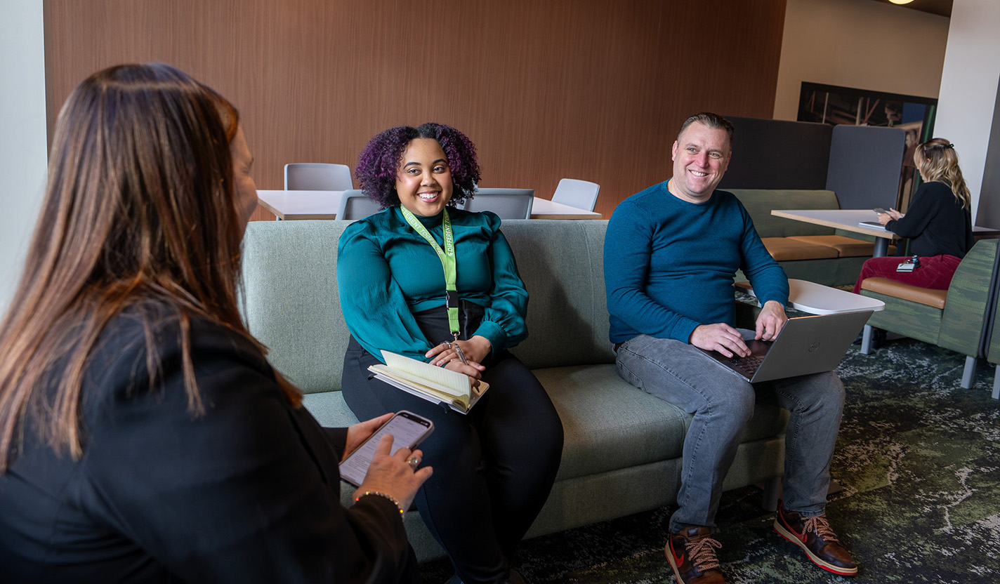Three people sit together, ta man am=nd a woman sit on a grey couch and a second woman sits in chair net to the couch while they talk and each hold a laptop, phone or paper notbookin their laps or hands.
