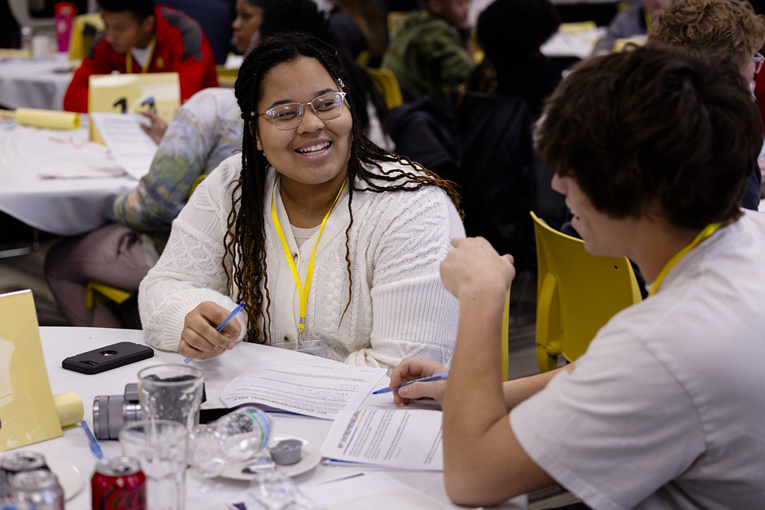 Two high school students talk to each other sitting at a table.
