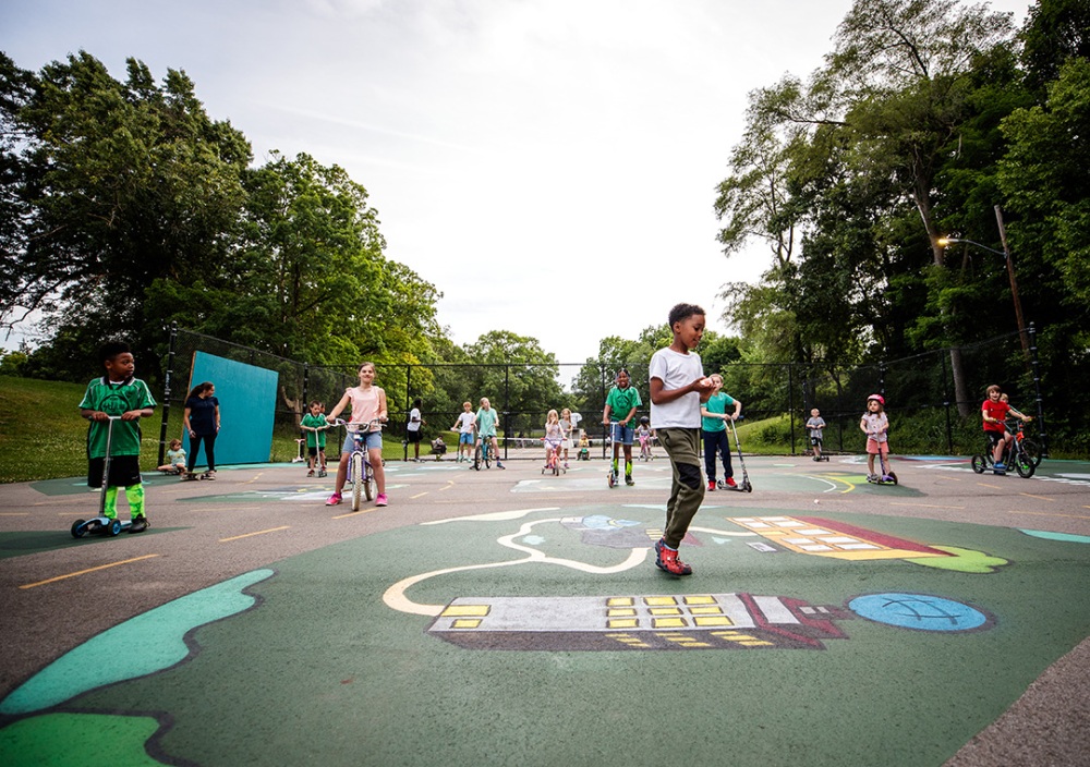 A large group of children from a neighborhood uses an old neighborhood converted tennis court as their playground and a place to ride their bikes and scooters. The cement is painted with several murals of the Flint area sights interconnected by two-way streets.