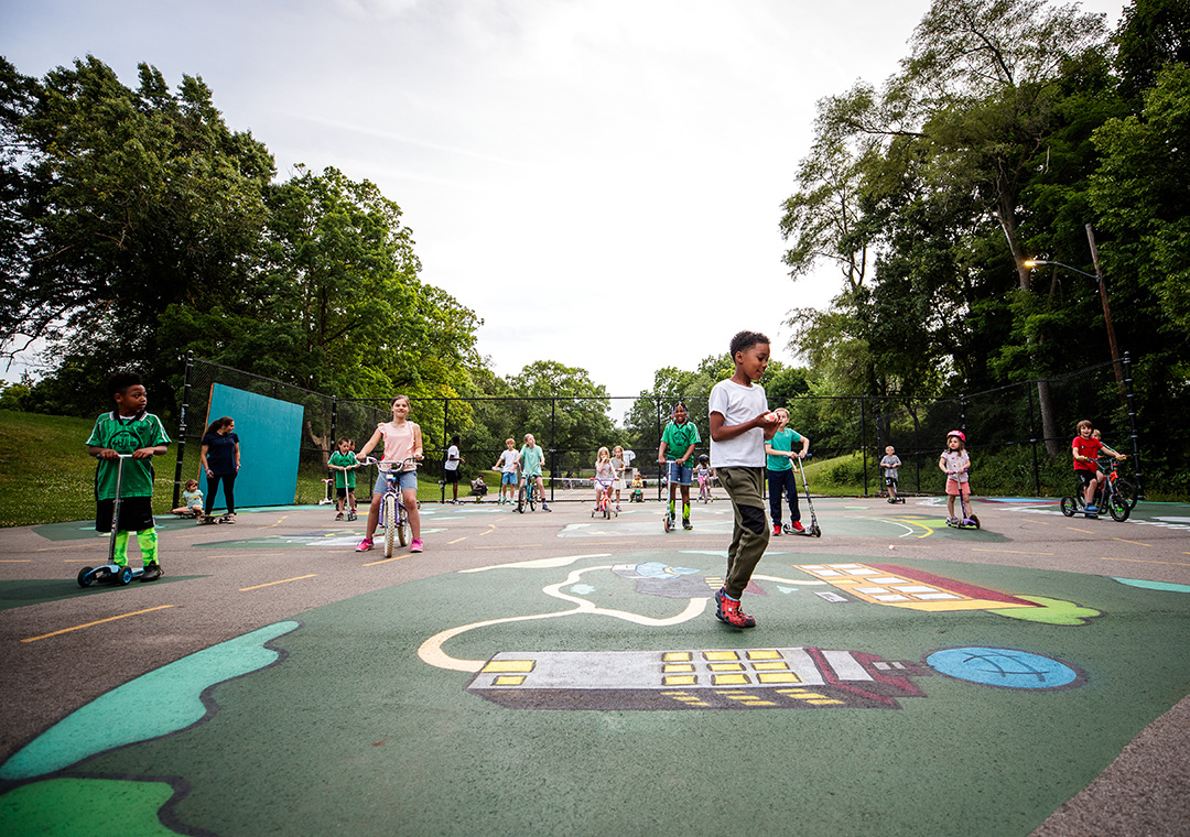 Un gran grupo de niños de un vecindario usa una antigua cancha de tenis convertida en patio de juegos y lugar para montar sus bicicletas y patinetas. El cemento está pintado con varios murales de los lugares de interés del área de Flint, conectados por calles de doble sentido.
