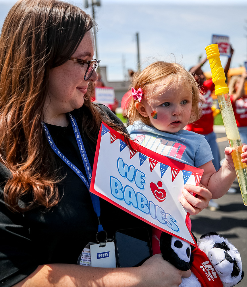A woman, holding a sign that reads, “We heart babies,” smiles down at an adorable infant she is holding in her arms.