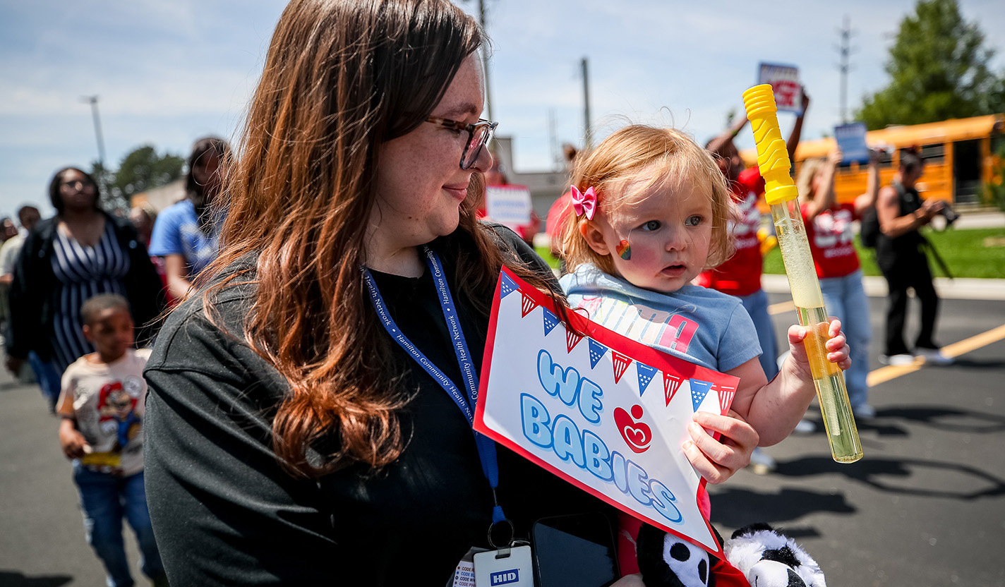 A woman, holding a sign that reads, “We heart babies,” smiles down at an infant she is holding in her arms.