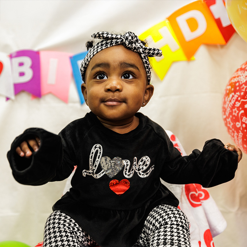 An adorable baby girl wears a shirt that says love while seated in front of birthday decorations.