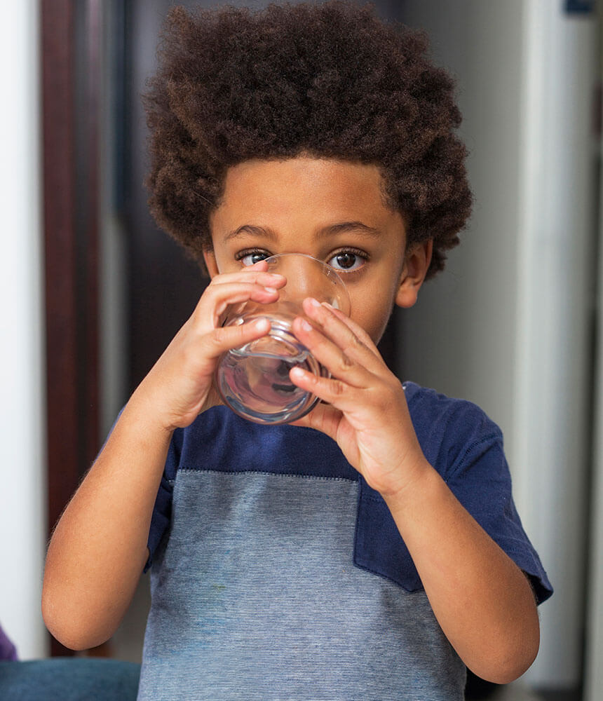 A young boy looks at the camera as he drinks water from a glass.