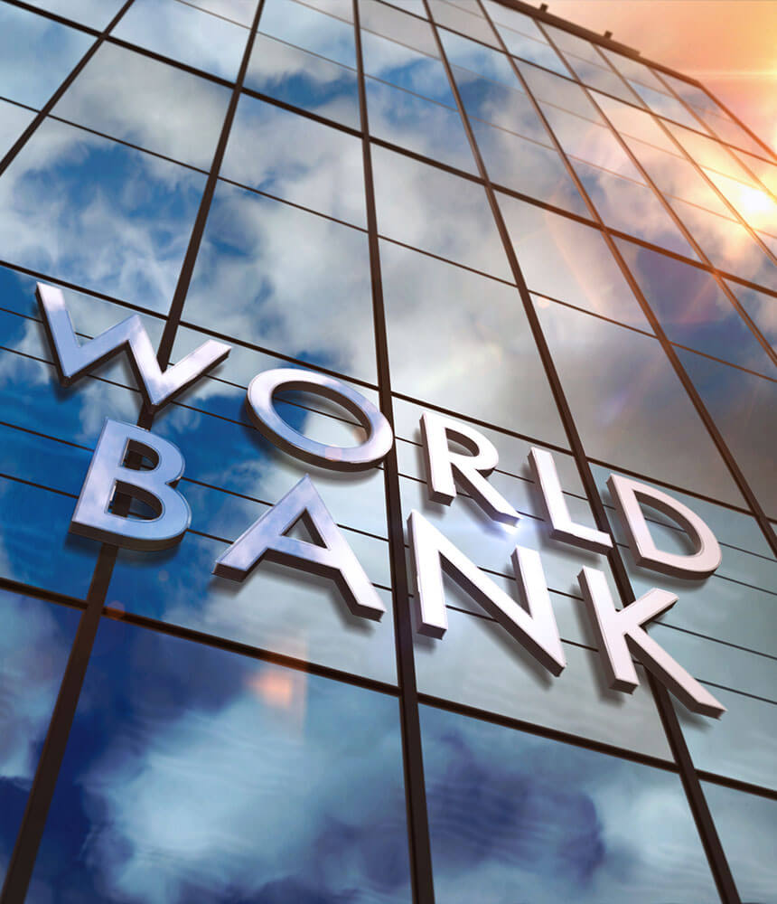 The glass facade of a building with letters spelling "World Bank." The glass facade of the building reflects the sun and sky.