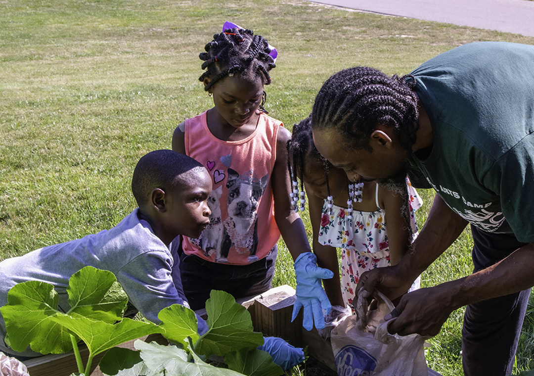 Un mentor adulto trabaja con tres jóvenes para mostrarles cómo plantar verduras en un jardín elevado en los terrenos de la Casa de Campo Berstom en Flint, Michigan.