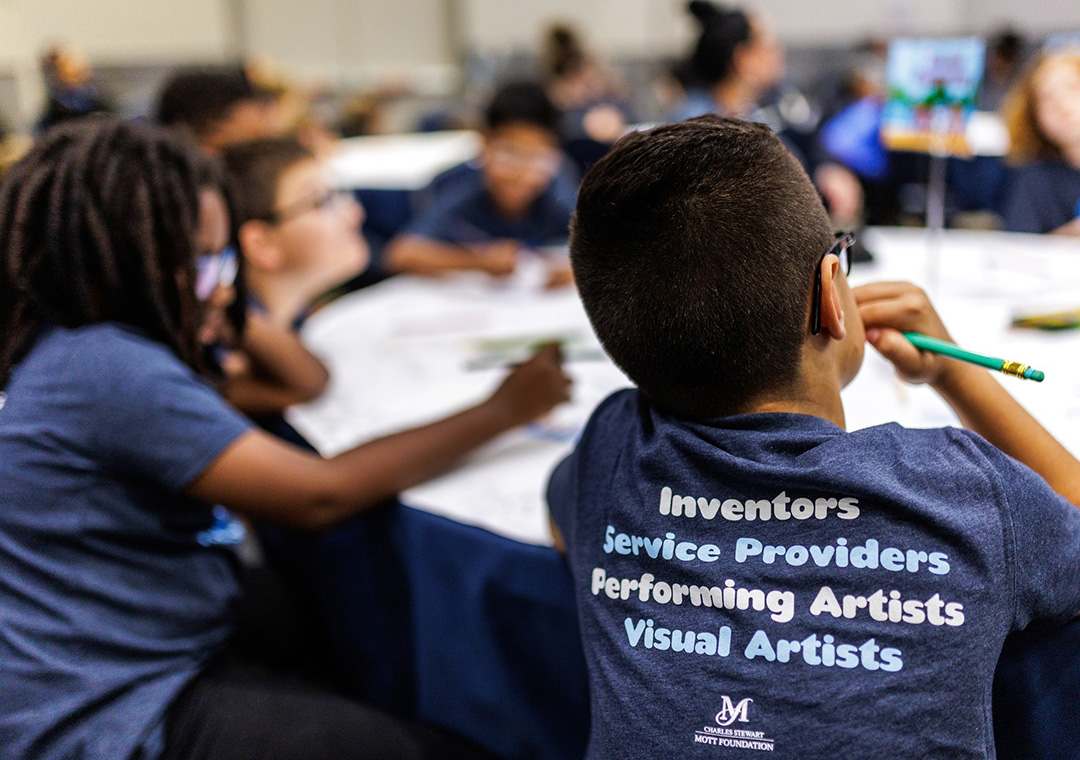 Two middle school-aged children, one a girl, the other a boy, sit at a table wearing matching dark blue t-shirts with the words "Innovators, Service Providers, Performing Artists, Visual Artists" and the Mott Foundation's previous logo on the back.