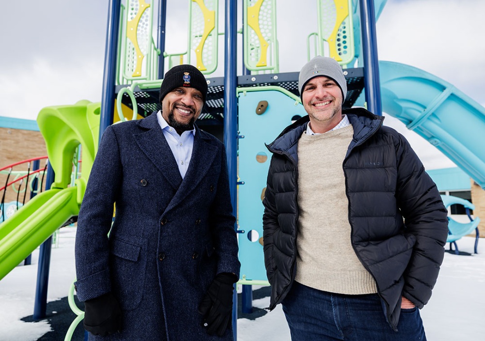 Two smiling men wearing winter coats and hats stand in front of colorful playground equipment.