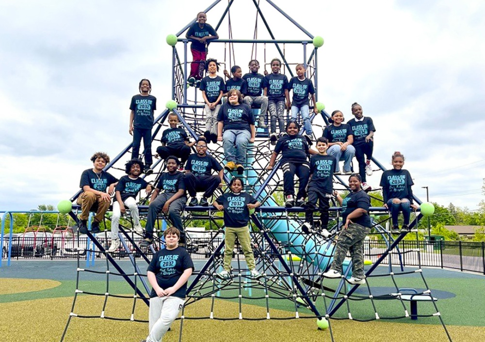 A group of middle school kids, all wearing dark blue t-shirts with the words Class of 2032, all sit on a newly built playground climbing dome made of crossbars and climbing rope nets as well as a yellow tube slide that is located behind them.