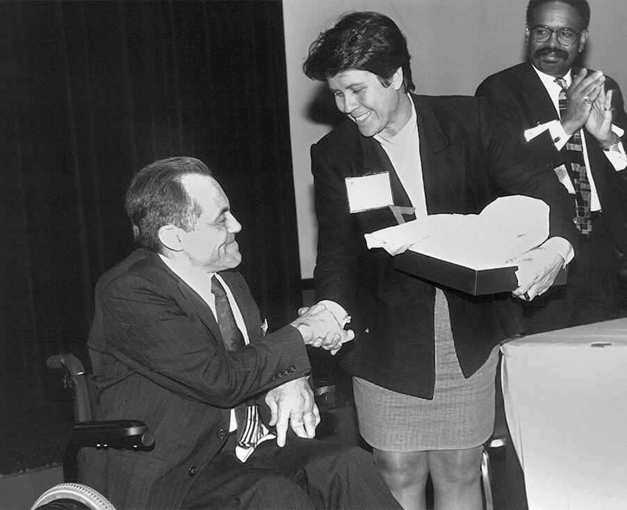 A black-and-white photo of a man seated in a chair and shaking hands with a woman who is standing next to him. The man and the woman are smiling at each other.