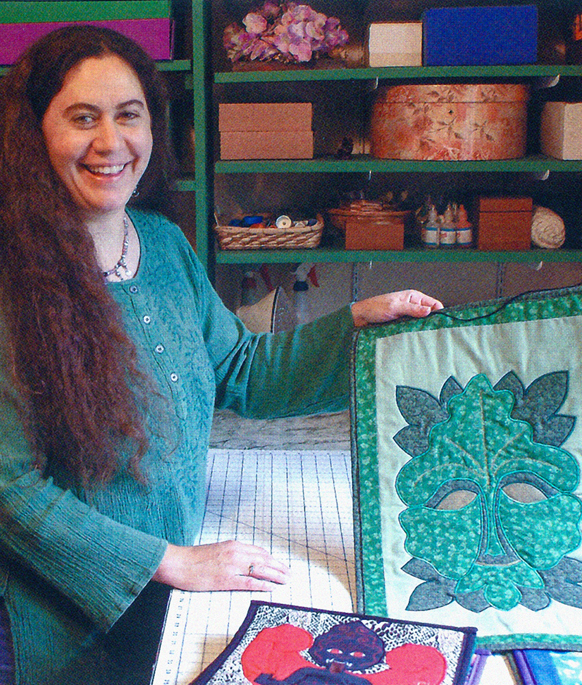 A photo of a smiling woman looking at the camera and holding up a quilt that she has made. The quilt’s design is that of large, abstract leaves on which is embroidered abstract features of a face. It is made in shades of green. The woman has long brown hair and is wearing a green blouse. On shelves behind her are various supplies for making quilts.