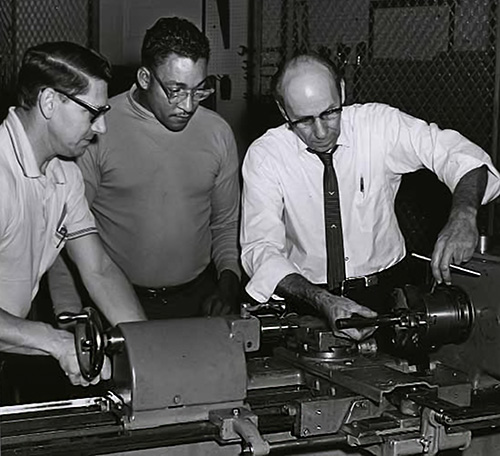 A black-and-white photo from the 1960s in which two men are watching an instructor show them how to operate a metal lathing machine.
