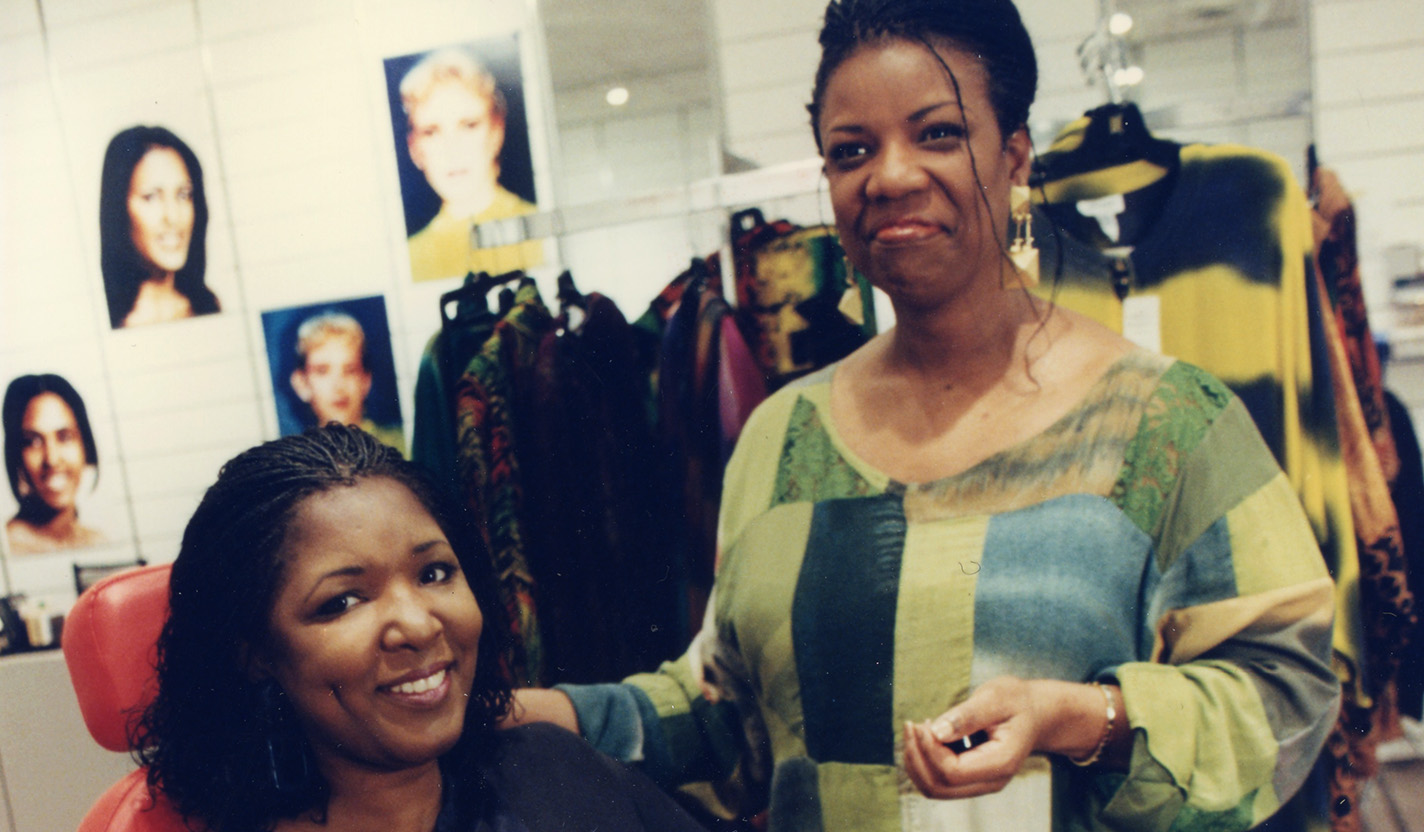 A photo of two women in a salon-like store. One woman is wearing a black blouse, seated in a red chair and is looking at and smiling at the camera. Standing next to her is the second woman, who is wearing a blouse of fabric featuring large blocks of green, blue and yellow colors. She is smiling and looking off to the left of the camera. Behind the two women is a rack of clothing and posters showing fashion models with various hairstyles.