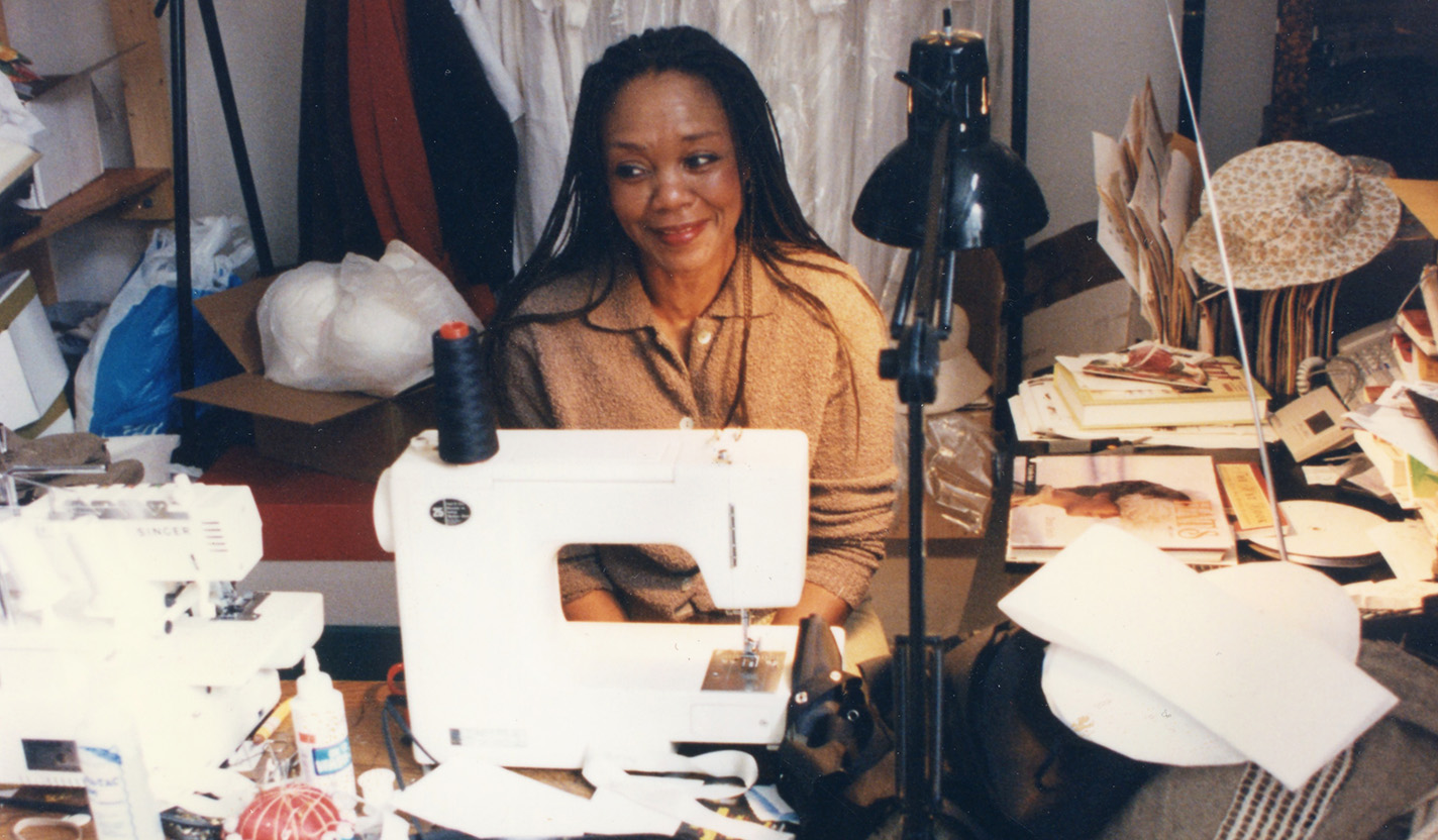 A photo of a woman wearing a light brown blouse and seated behind a white sewing machine. She is smiling and looking off to the side of the camera. Around her are stacks of papers and sewing supplies, and behind her is a rack on which clothing is hung.