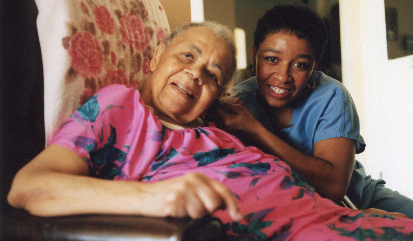 A photo of two women who are smiling and looking at the camera. The woman on the left is perhaps in her seventies and is sitting in a recliner-type chair. She is wearing a bright pink dress with blue flowers in the design. The woman on the right is in her thirties and is wearing a blue medical smock and pants.