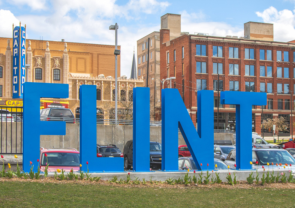 An image of the large iron FLINT sign painted in sky blue with the historic facade of the Capitol Theatre and the Dryden Building in the background, and fresh tulips growing at its base as spring comes into full bloom.
