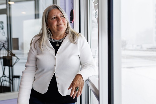 A woman with blond hair who is wearing a dark dress and white cardigan leans against a wall of windows as she watches snow fall. She has a slight smile on her face.