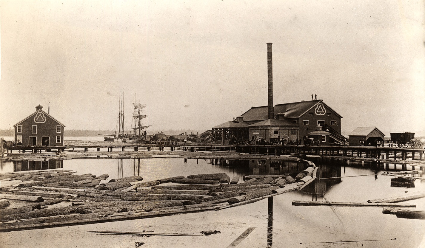 L.G. Mason sawmill, foot of Fourth Street on Muskegon Lake, Muskegon, Michigan, circa 1865-1878. There is a log boom in the foreground, a schooner in the background, and a tramway connects the sawmill to another building.