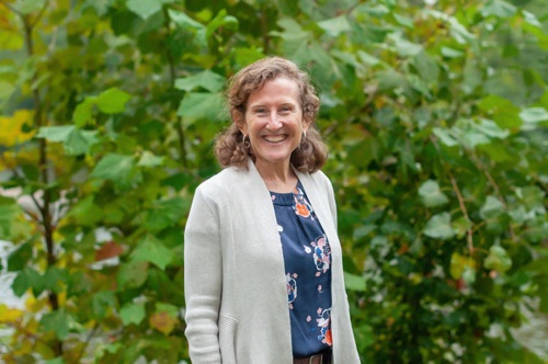 A smiling white woman with light brown hair wears a blue top with a colorful pattern and a light gray open sweater. She is standing in front of young, leafy green trees that are just showing the first signs of yellow and orange fall colors.