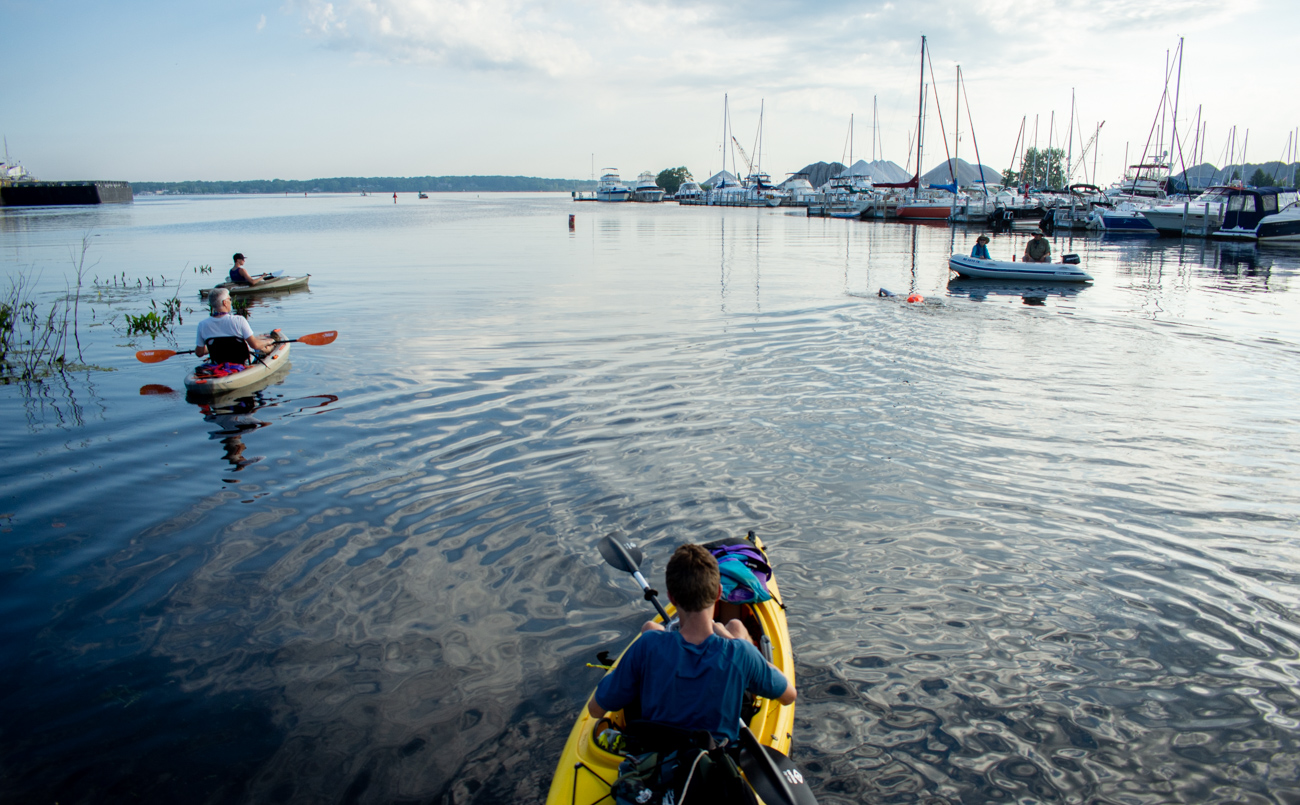 Three people in single kayaks and two people in a small inflatable boat have their backs to the camera as they float to the side and behind a swimmer wearing an orange bathing cap in Muskegon Lake. There are ripples on the water and a few clouds in a blue sky. There are sailboats moored in the top right of the photo.