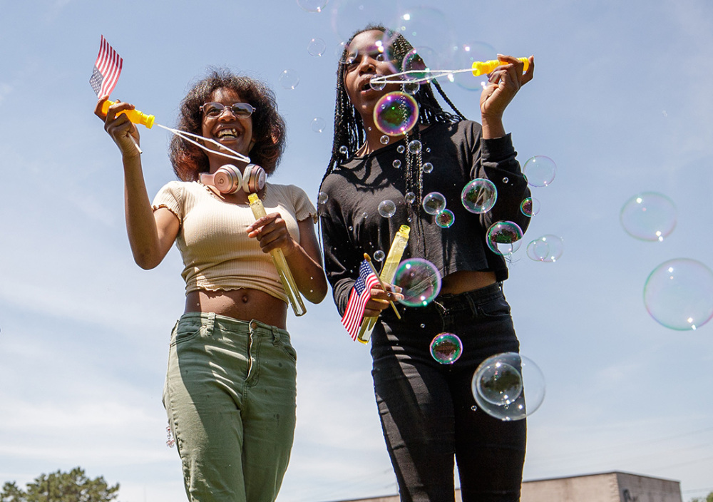 Two smiling girls holding small American flags blow bubbles on a sunny day.