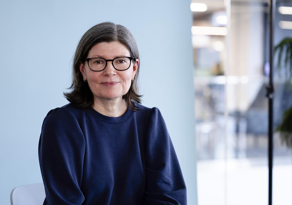 A woman with glasses sits facing the camera against a light blue backdrop, photographed indoors with a softly blurred library environment in the background.