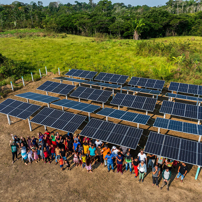 A large group of adults and children stand under a row of solar panels that are part of an array in a forest area.