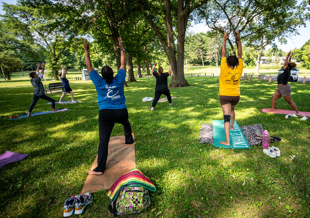Six people, dressed in exercise clothes, are doing a sun salutation yoga strech while on their yoga mats in Ballinger Park in Flint, Michigan.