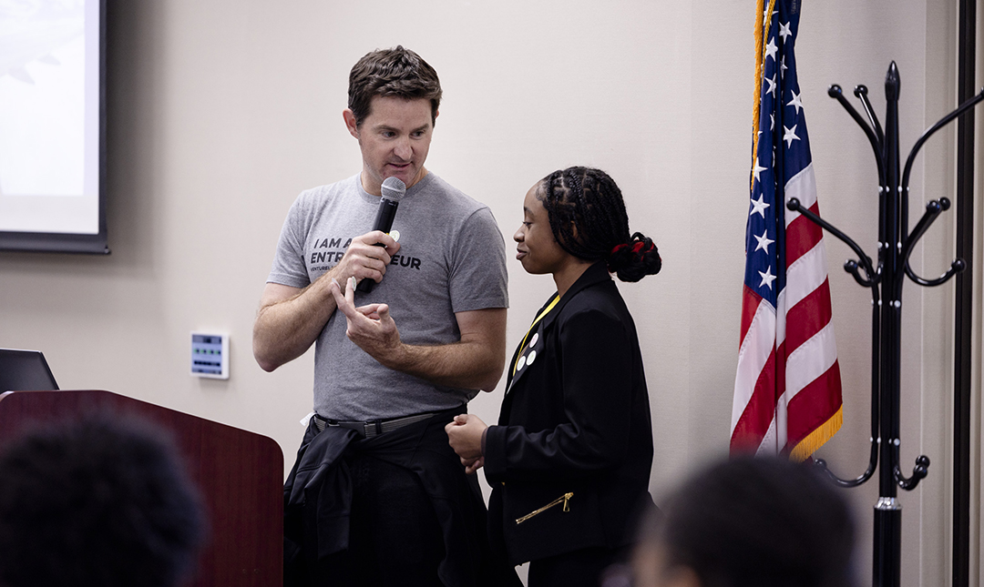 An adult speaks to a student using a microphone. The adult’s shirt reads “I am an entrepreneur.” An American flag and a podium are visible.