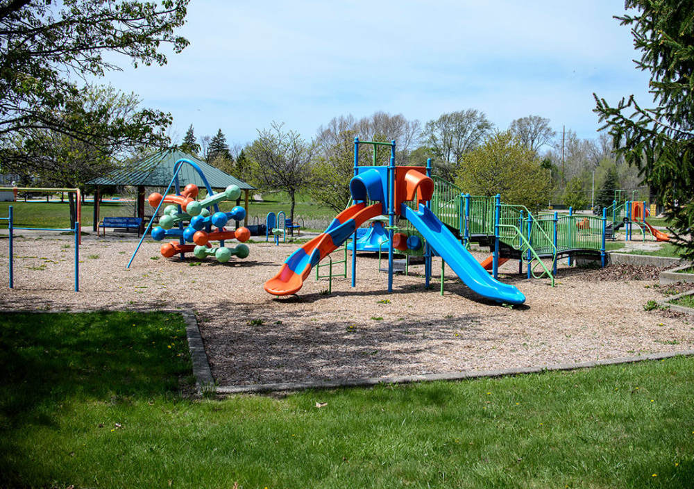 A playground with bright blue and orange slides and connected platforms sits on gravel, with trees and grassy areas surrounding the play space.