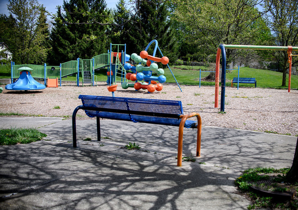 A blue metal bench sits on a concrete pad in the foreground, facing an empty playground with slides, climbing structures, swings, and gravel surfacing under leafy trees.