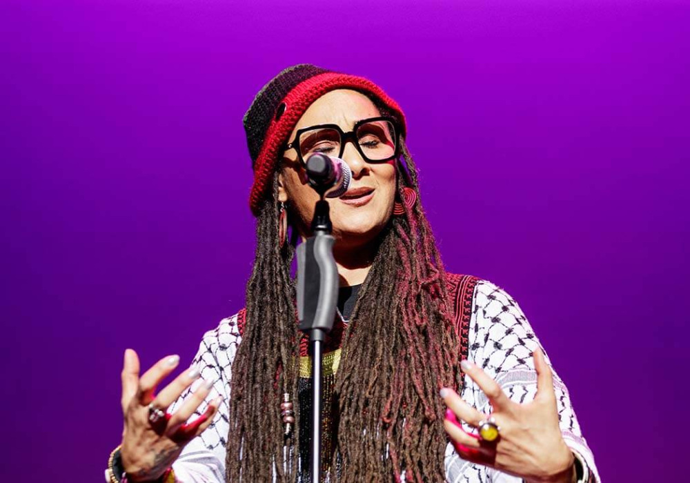 Person with long dreadlocks wearing large glasses, a red knit hat, and a patterned black-and-white shawl stands at a microphone onstage, gesturing with both hands against a purple-lit background.