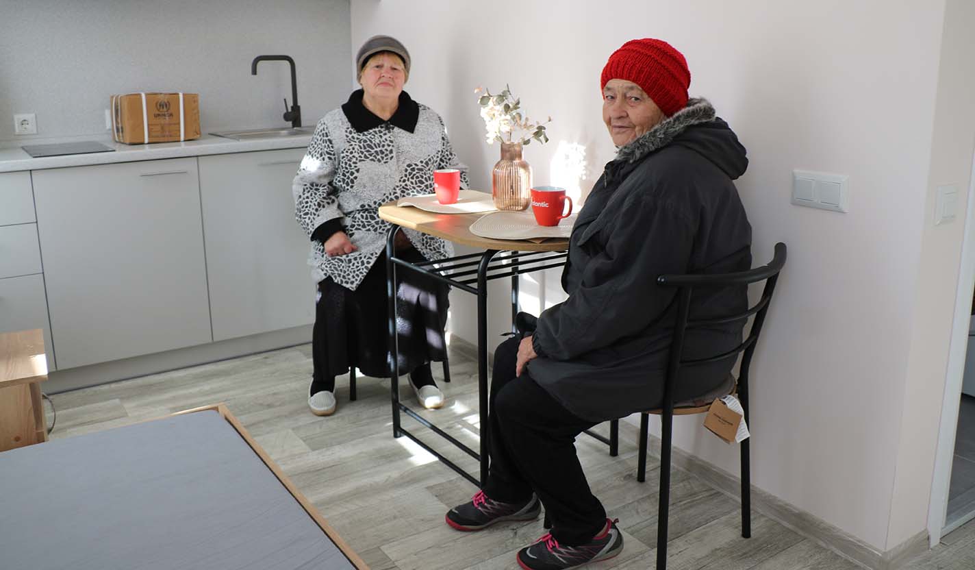 Two older adults sit at a small table with cups in a bright kitchen inside community foundation-supported housing for individuals receiving humanitarian relief assistance related to the war in Ukraine.