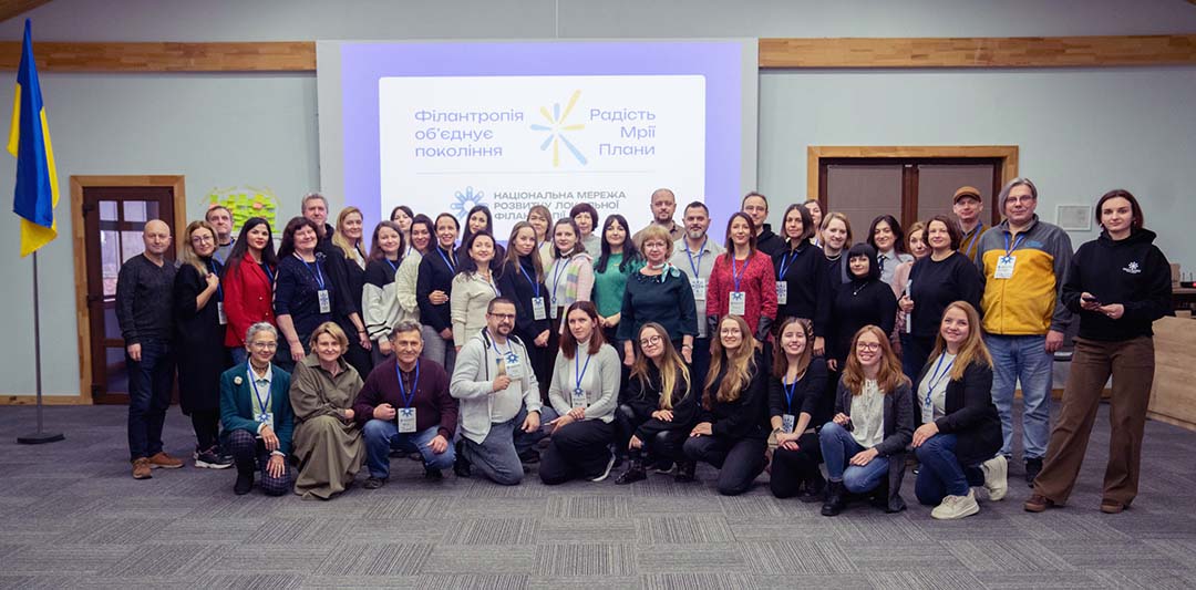 Large group of community foundation partners and participants pose together in front of a presentation screen and Ukrainian flag, representing collaboration and network-building supporting humanitarian relief efforts related to the war in Ukraine.