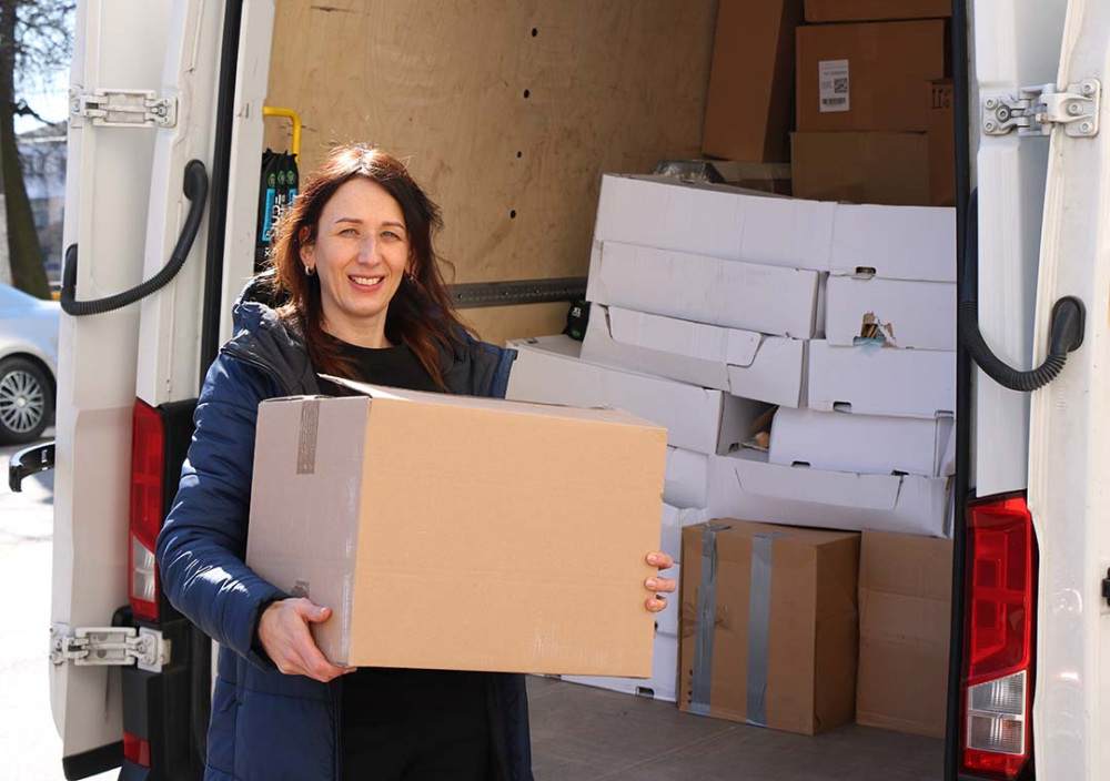 Community foundation representative holds a cardboard box while distributing humanitarian relief supplies from a van filled with packages for community support related to the war in Ukraine.