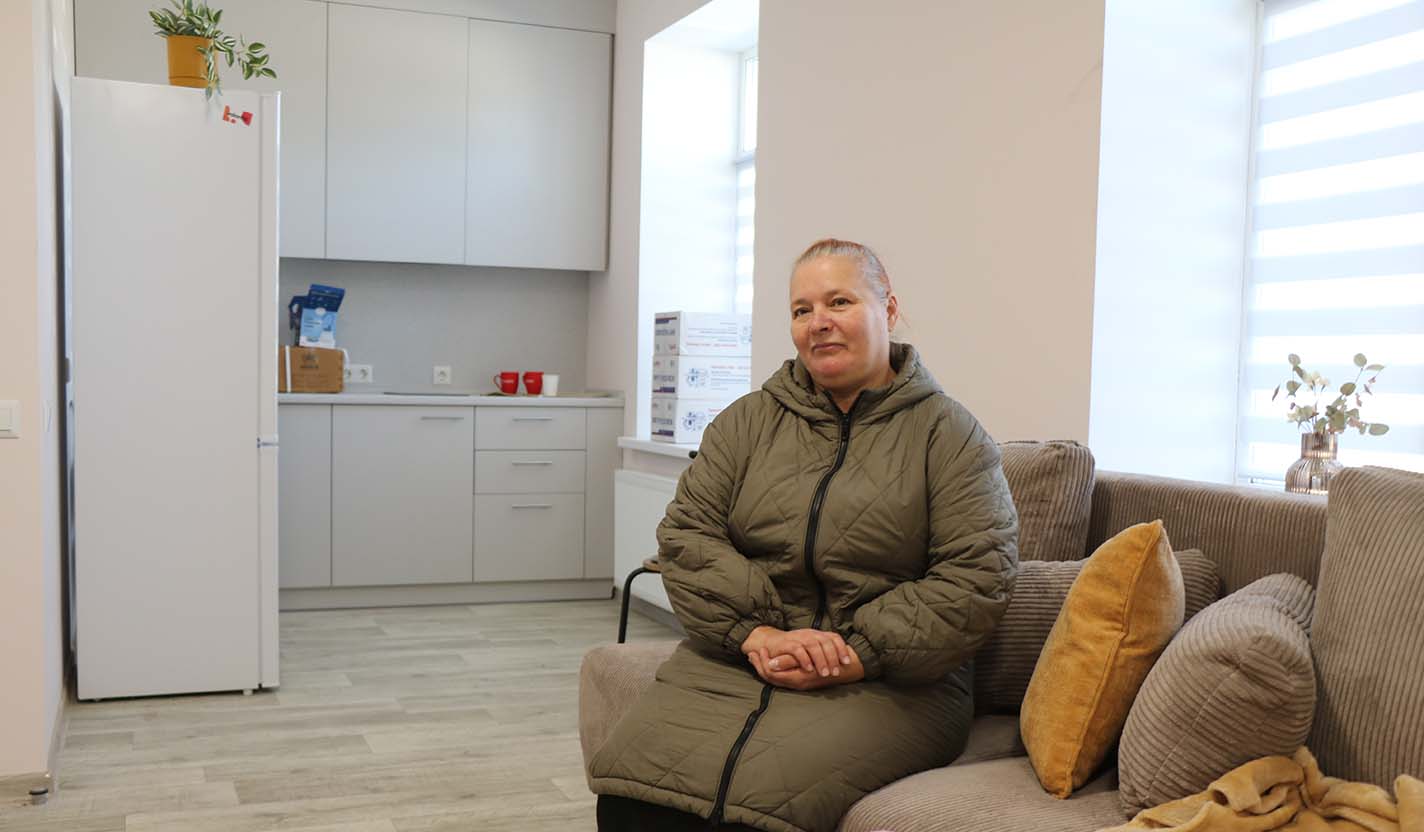Individual sits on a couch in a bright, renovated apartment with a kitchenette, representing safe and stable living conditions provided through community foundation-supported housing for individuals receiving humanitarian relief assistance related to the war in Ukraine.