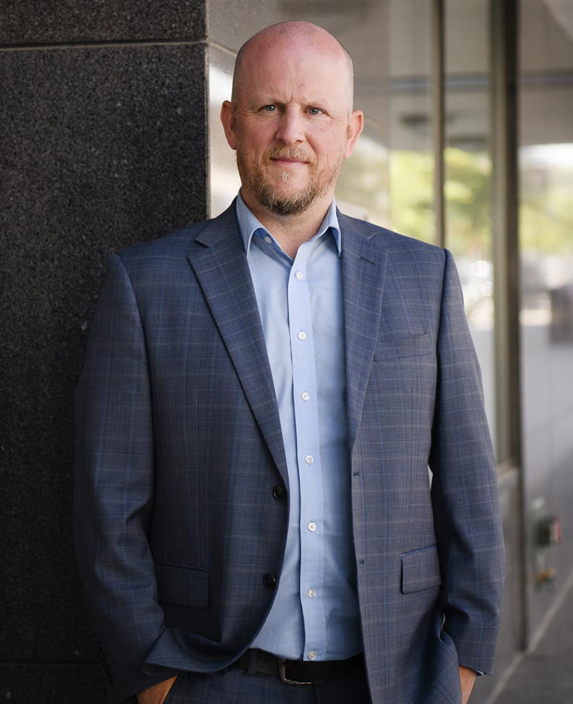 Person wearing a suit jacket stands outdoors beside a building and looks toward the camera, with glass windows and architectural elements in the background.