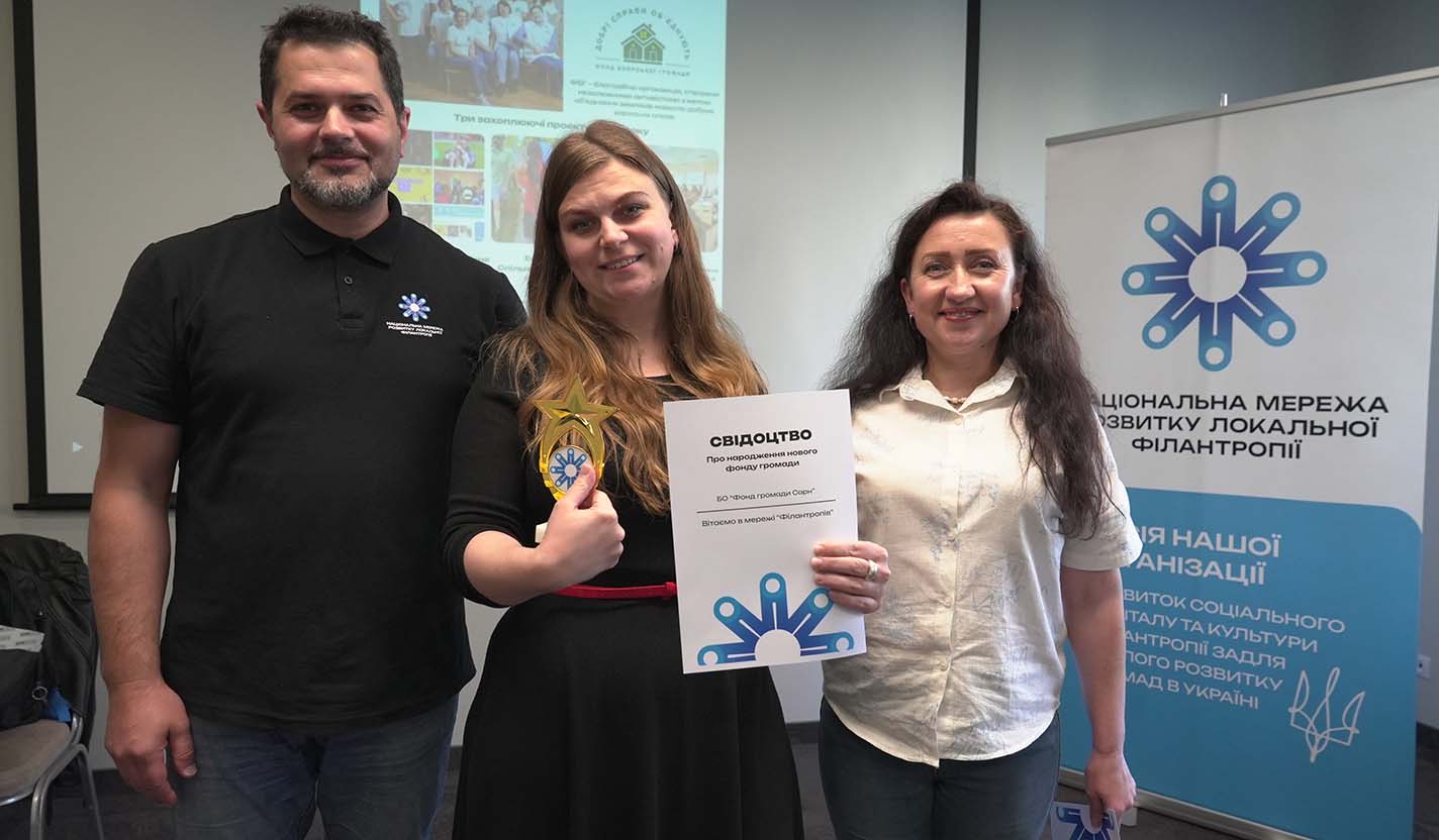 Three individuals stand together indoors holding a certificate and award in front of a community foundation network banner, representing recognition and collaboration supporting humanitarian relief efforts related to the war in Ukraine.